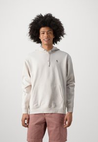 Helles beiges Hoodie aus weichem Stoff, ausgestattet mit einer Vordertasche, einer Kapuze mit Kordelzug und einem kleinen Logo auf der Brust. Kombiniert mit rosa Shorts.