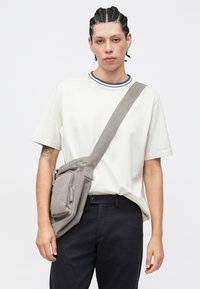 Grå crossbody-taske med tekstureret finish, lynlåslomme foran, båret af en person iført en hvid skjorte med blåstribet halsudskæring.