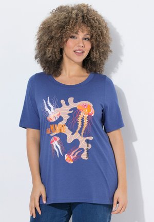 Frau mit lockigem Haar, die ein blaues T-Shirt mit bunten Quallen- und Korallengrafiken trägt, kombiniert mit blauen Jeans, stehend vor einer weißen Wand.