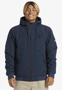 Marineblaue Kapuzenjacke mit einem Reißverschluss vorne, elastischen Bündchen und zwei Seitentaschen. Hergestellt aus einem glatten, leichten Material.