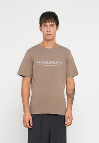 Καφέ βαμβακερό μπλουζάκι με κοντά μανίκια, στρογγυλή λαιμόκοψη, με λευκό λογότυπο στο κέντρο του στήθους. Λιτή υφή, άνετη εφαρμογή.