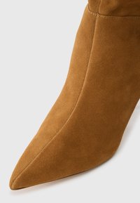 Bottine à talons aiguilles en daim marron, avec un bout pointu, présentant une texture lisse et des détails cousus le long de la couture.