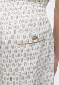 Tissu blanc avec des motifs géométriques marron composés de formes rectangulaires et de cercles, une poche avec un petit logo brodé et un bras visible sur le côté.