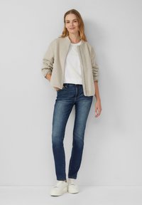 Veste bomber beige, T-shirt blanc, jeans bleu foncé et baskets plateformes blanches. Texture lisse, coupe décontractée, sans motifs évidents.