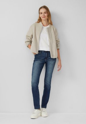 Veste bomber beige, T-shirt blanc, jeans bleu foncé et baskets plateformes blanches. Texture lisse, coupe décontractée, sans motifs évidents.