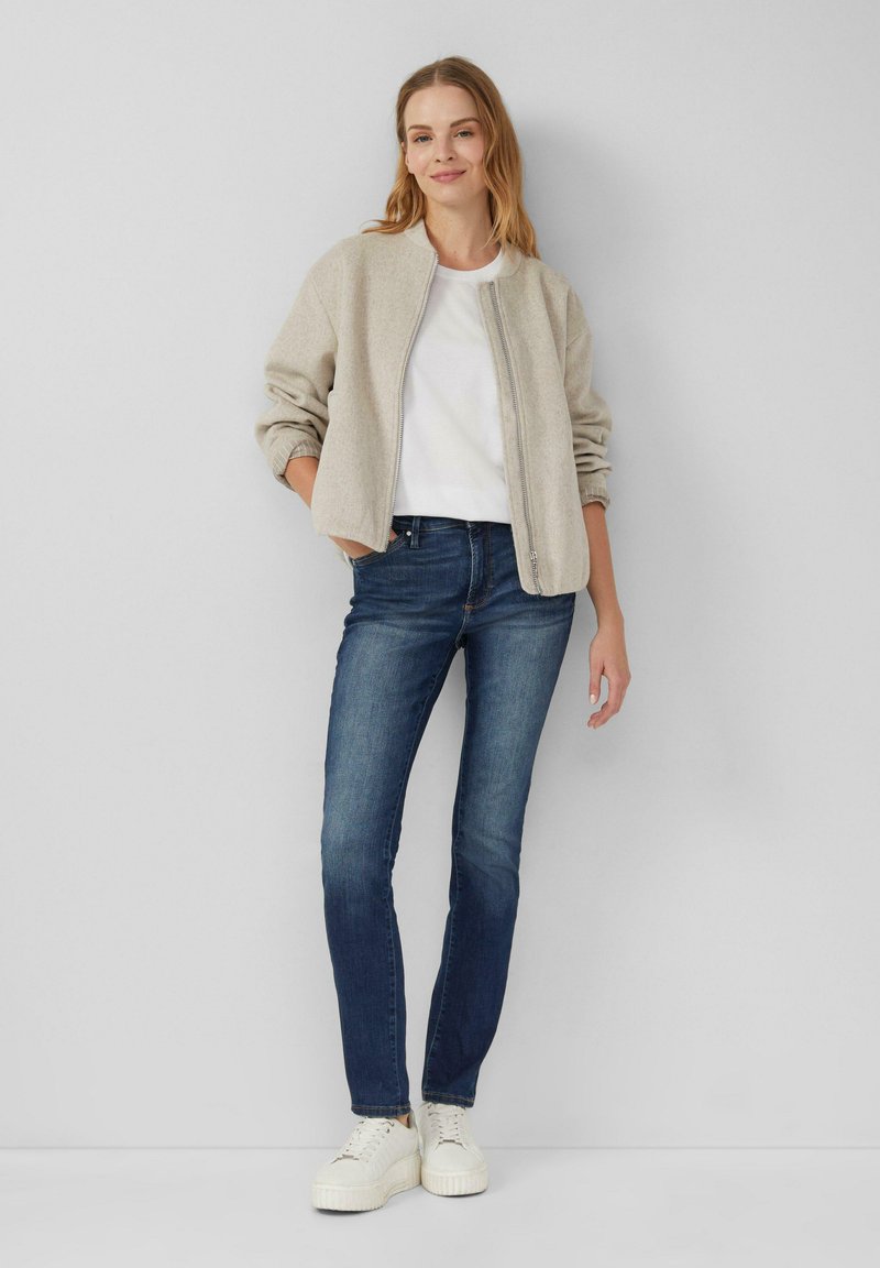Veste bomber beige, T-shirt blanc, jeans bleu foncé et baskets plateformes blanches. Texture lisse, coupe décontractée, sans motifs évidents.