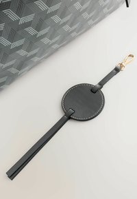 Šedá taška z plátna so vzorom a tmavou koženou značkou. Značka má okrúhly tvar, zlatú kovovú výbavu a tenký remienok. Výrazné prešívanie.