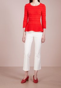 Femme portant un haut péplum rouge texturé, un pantalon blanc court et des chaussures à talons carrés rouges tissées, debout devant un fond beige.