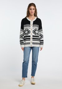 Karierte schwarze und weiße Strickjacke mit rundem Hals, Knöpfen vorne und langen Ärmeln, kombiniert mit hellblauen Jeans und weißen Sneakers.