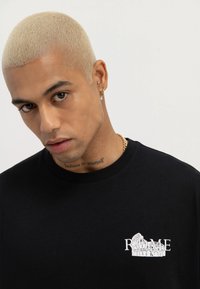 Uomo con capelli biondi corti che indossa una T-shirt nera con la scritta "ROME" e una grafica del Colosseo, con una catena d'oro, che guarda leggermente verso il basso.