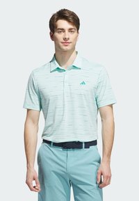 Polo shirt a maniche corte di un verde acqua chiaro con una texture a motivo e il logo Adidas. Indossato con pantaloni chiari coordinati e una cintura blu navy.