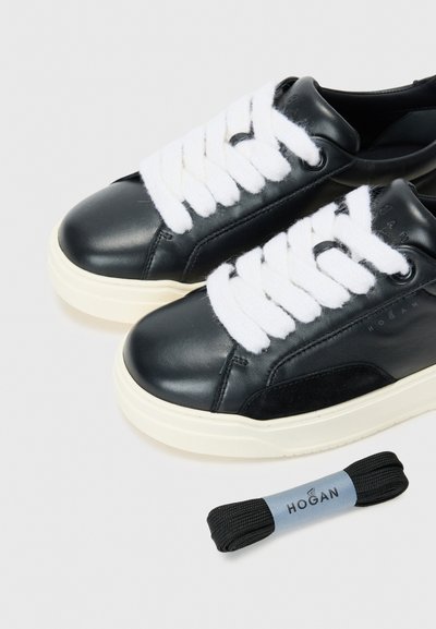 Black leather sneakers with white plush laces and a cream rubber sole. A pair features a suede detail and a replacement black lace nearby.