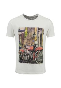 Camiseta blanca de hombre con un gráfico vintage de motocicleta roja, situado frente a un fondo urbano.