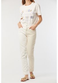 T-shirt blanc avec du texte multicolore, pantalon crème taille haute et sandales transparentes avec des accents dorés. Tissu lisse, design ajusté.