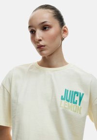 Cremefarvet bomulds T-shirt med korte ærmer og rund halsudskæring, med "JUICY LEMON" trykt i teal og gradient gul skrifttype.