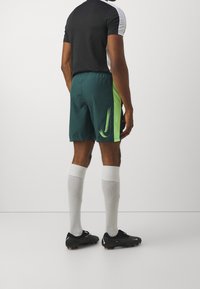 Hombre vistiendo una camiseta deportiva blanca y negra, pantalones cortos verde oscuro con una franja y un logotipo en verde neón, calcetas blancas hasta la rodilla y botines de fútbol negros.