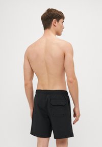 Sorte svømmeshorts med teksturert overflate, elastisk midjebånd og baklomme. Modell sett bakfra, bar hud synlig.