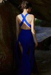 Modré šaty s priliehavým telom a odhaleným chrbtom, so skríženými ramienkami a voľnou sukňou s vysokými rozparkami po oboch stranách.