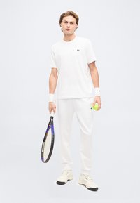 Νεαρός άνδρας με λευκή τενιστική στολή κρατώντας ρακέτα και μπάλα, στέκεται μπροστά σε απλό φωτεινό φόντο.