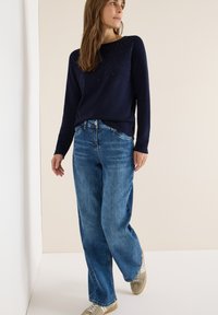 Dunkelblauer Pullover mit langen Ärmeln, verziert mit verstreuten Strasssteinen auf der Vorderseite, kombiniert mit lässigen hellblauen Jeans und beigen Sneakers.