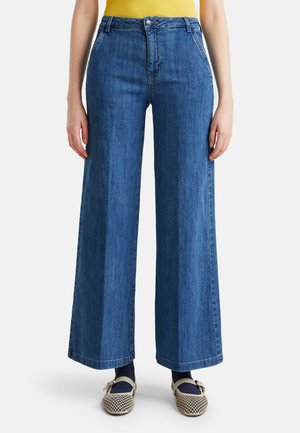 Blaue Jeans mit weitem Bein, getragen mit navyblauen Socken und beigen gewebten Mary Jane Schuhen, kombiniert mit einem gelben Top.