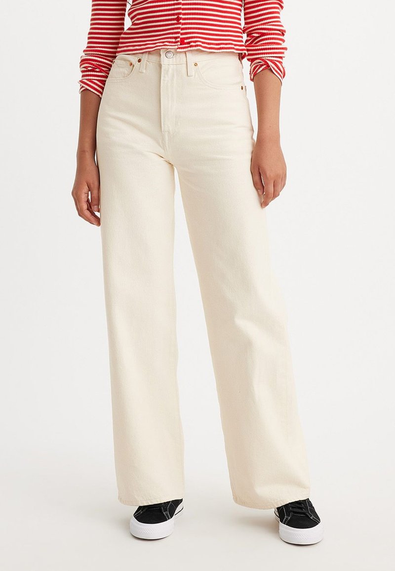 Jeans taille haute, à jambes larges en crème, en coton. Dotés de cinq poches et d'une fermeture à bouton, associés à des baskets noires.