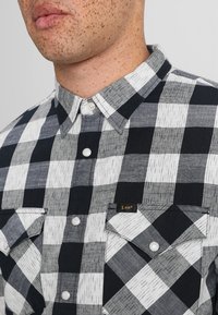 Camicia a scacchi bianca e nera con colletto corto, chiusura a bottoni frontale, due taschini sul petto e etichetta con logo su una tasca. Materiale in misto cotone.