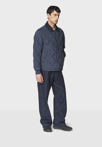 Quiltet navy pufferjakke med krave, to lommer og ribbede manchetter, parret med mørke vidde jeans og sort fodtøj.
