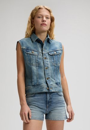 Νεαρή γυναίκα με κυματιστά ξανθά μαλλιά, φοράει αμάνικο μπλε ντένιμ γιλέκο και ταιριαστά μπλε σορτς ντένιμ, μπροστά σε ένα απλό φόντο.