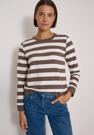 Femme aux cheveux bruns courts portant un t-shirt à manches longues rayé marron et blanc et un jean bleu, debout avec une main dans la poche.