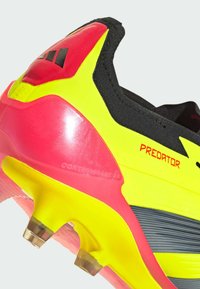 Scarpini da calcio neri, gialli e rosa con una tomaia sintetica liscia, un design del tallone prominente e tacchetti trasparenti su una suola texturizzata.