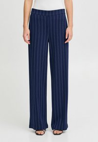 Pantalons larges bleu marine en tissu doux avec des rayures blanches verticales et une taille plate. Portés avec des sandales noires.