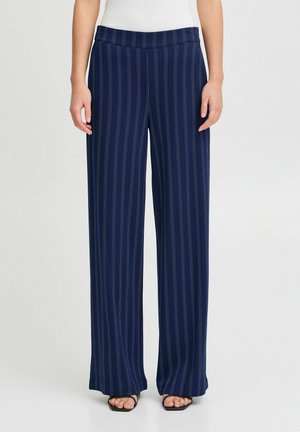 Pantalons larges bleu marine en tissu doux avec des rayures blanches verticales et une taille plate. Portés avec des sandales noires.