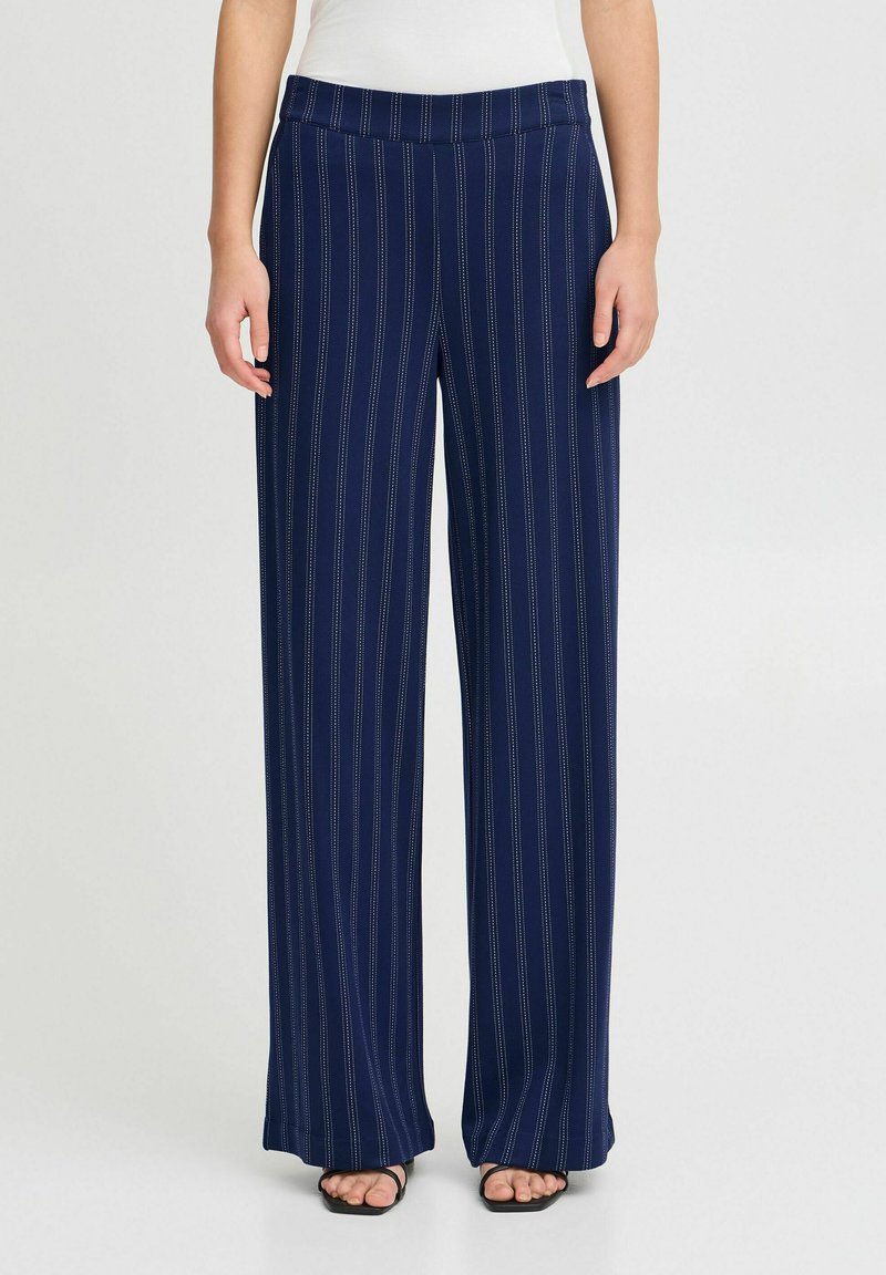 Pantalons larges bleu marine en tissu doux avec des rayures blanches verticales et une taille plate. Portés avec des sandales noires.