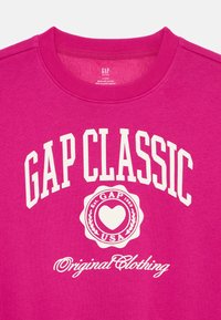 Magenta színű pulóver, melyen a "GAP CLASSIC" felirat és egy kör alakú logó látható, rajta "Est. GAP 1969" és "USA" felirat fehér színben. Pamut anyagú, kerek nyakkivágású.