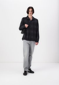 Svart rutete skjorte, grå denimjeans, svarte joggesko med hvite striper, holder en liten svart veske, står mot en hvit bakgrunn.