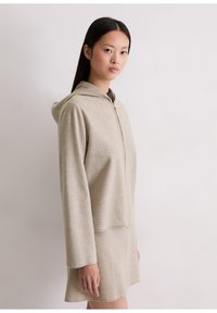 Beige hooded top met een rits aan de voorkant, zachte textuur, lange mouwen, gecombineerd met een bijpassende knielange rok in een vergelijkbare kleur en stof.
