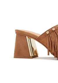 Sandale à talon en daim marron avec des détails en franges, accents métalliques dorés sur le talon et une semelle intérieure beige lisse.