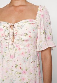 Vestido floral con mangas abullonadas cortas, con un fondo pastel claro, flores en tonos rosa y verde, y un detalle de escote con cordón.