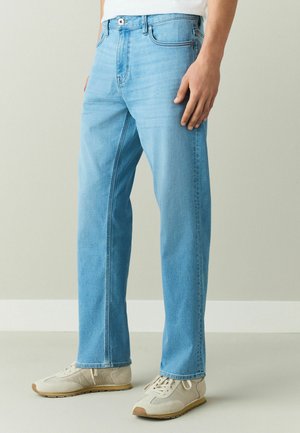 Mann trägt hellblaue Straight-Leg-Jeans und beige Sneaker, steht mit einer Hand entspannt an der Seite.