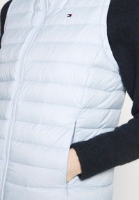 Personne portant un gilet matelassé bleu clair avec une fermeture éclair à l'avant et la main dans la poche latérale, par-dessus un pull à manches longues foncé.