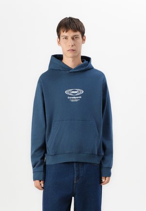 Jonge man draagt een blauwe CoreSport© hoodie met een voorzak en donkerblauwe jeans, staand tegen een effen witte achtergrond.