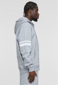 Grå hoodie med hvide striber på ærmerne, kængurulomme og lynlås. Stoffet ser blødt og tekstureret ud, med en afslappet pasform og hætte.