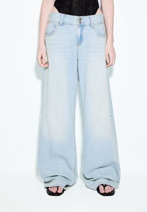 Osoba nosiaca veľmi široké svetlomodré džínsy v kombinácii s čiernymi žabkami a čiernym čipkovaným topom, zobrazená od pása po nohy.