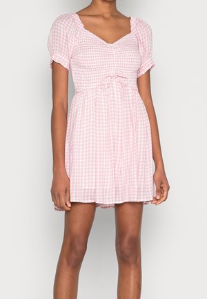 Kobieta ubrana w krótką różowo-białą sukienkę w kratkę gingham z bufkami na rękawach i talią z małą kokardką.
