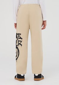 Beżowe dresy z elastycznym pasem, z dużym czarnym nadrukiem w stylu graffiti na lewej nodze. Gładka bawełniana tekstura, luźny krój.