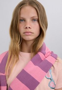 Bufanda a rayas en algodón rosa y morado, colocada sobre una camiseta rosa, con un parche en forma de corazón que presenta elementos decorativos.
