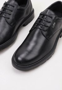 Chaussures en cuir noir avec une finition lisse, bouts arrondis, six œillets avec lacets et semelles en caoutchouc texturé. Aucune motif ou accent visible.