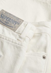 Jeans de mezclilla blanca con una superficie texturizada, una etiqueta de cuero gris con la marca "Superdry" y un pequeño detalle de botón metálico.