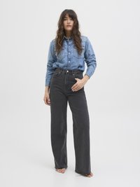 Kvinne med langt bølgete hår iført en blå denimskjorte som er stumpet inn i høytlivede, svarte vide bukser, stående barføtt mot en hvit bakgrunn.